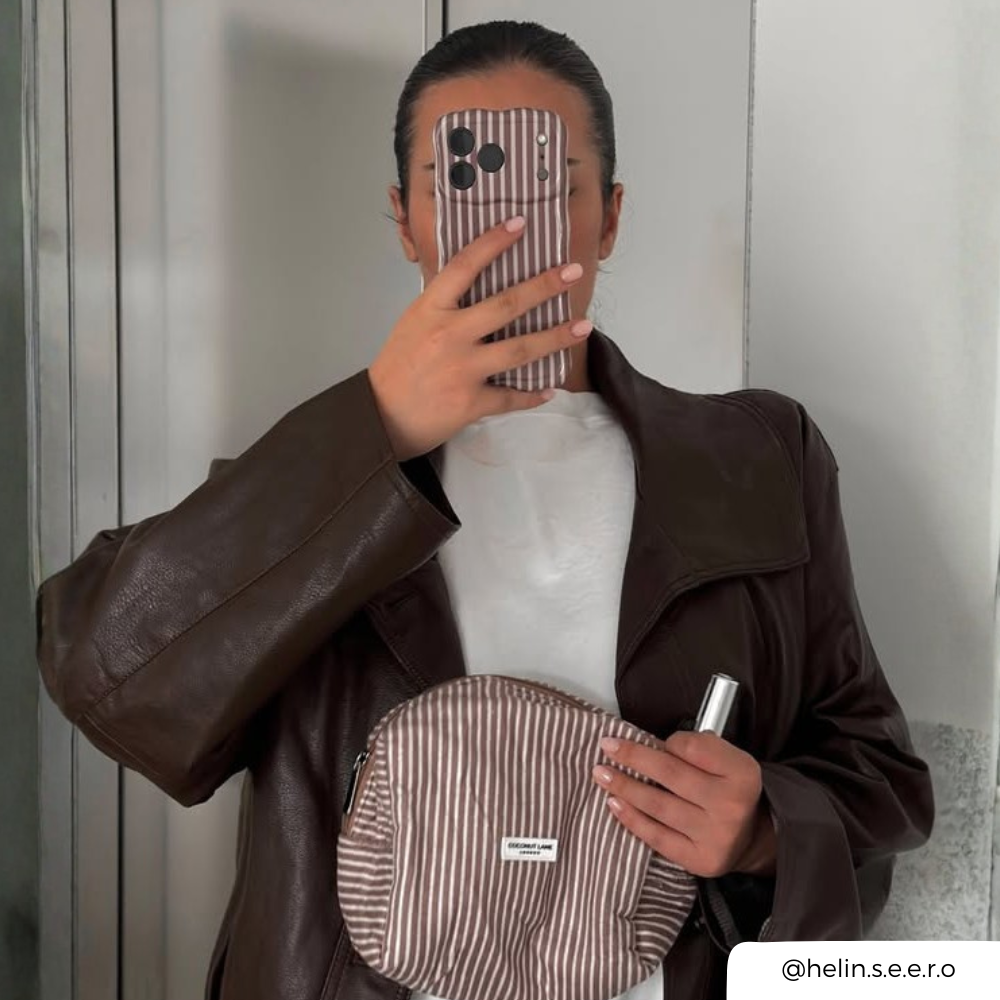 A woman wearing a dark brown faux-leather jacket over a white t-shirt and blue jeans, holding a brown striped phone case, taking a mirror selfie.. She is also holding a matching makeup bag.