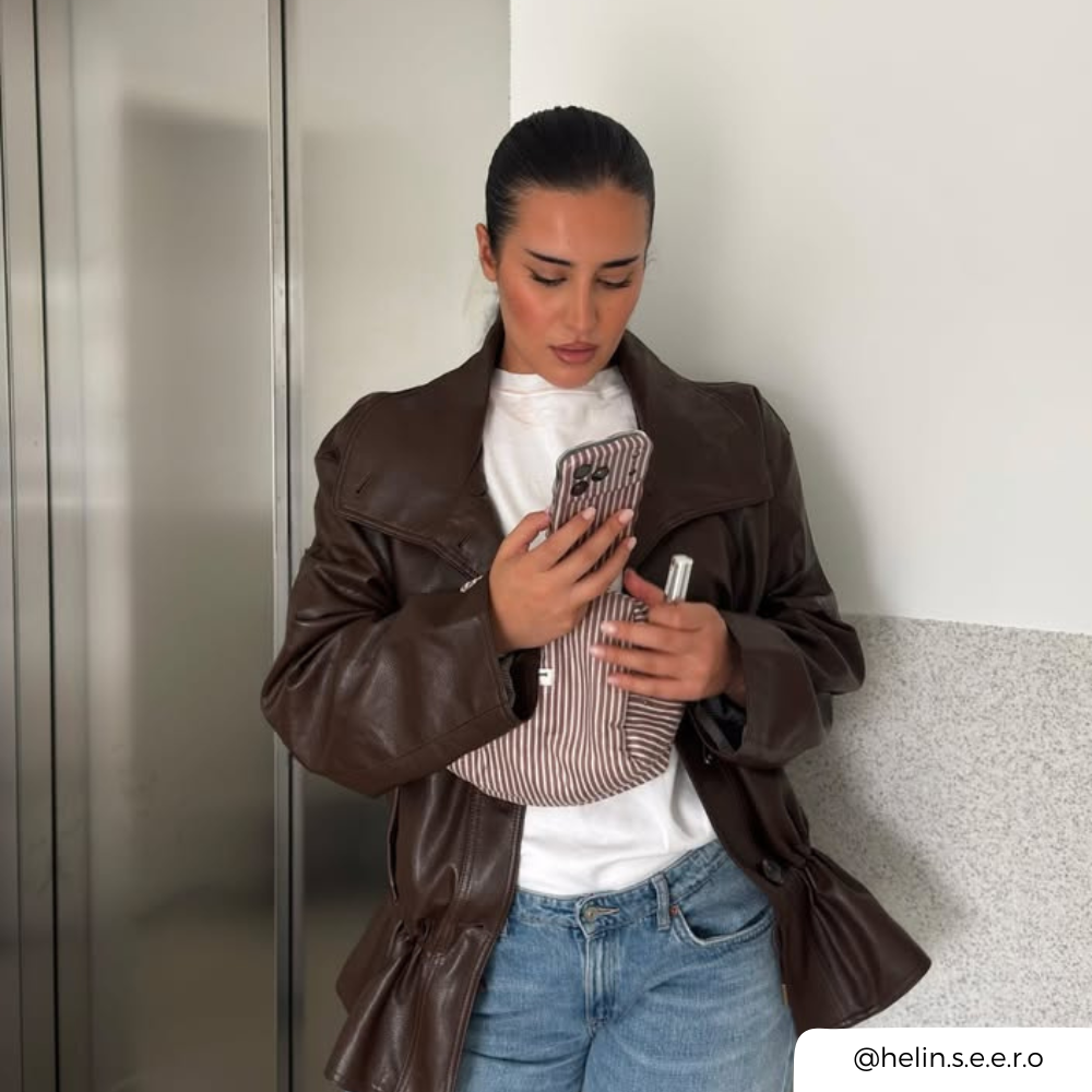 A woman wearing a dark brown faux-leather jacket over a white t-shirt and blue jeans, holding a brown striped phone case in front of her face. She is also holding a matching makeup bag.