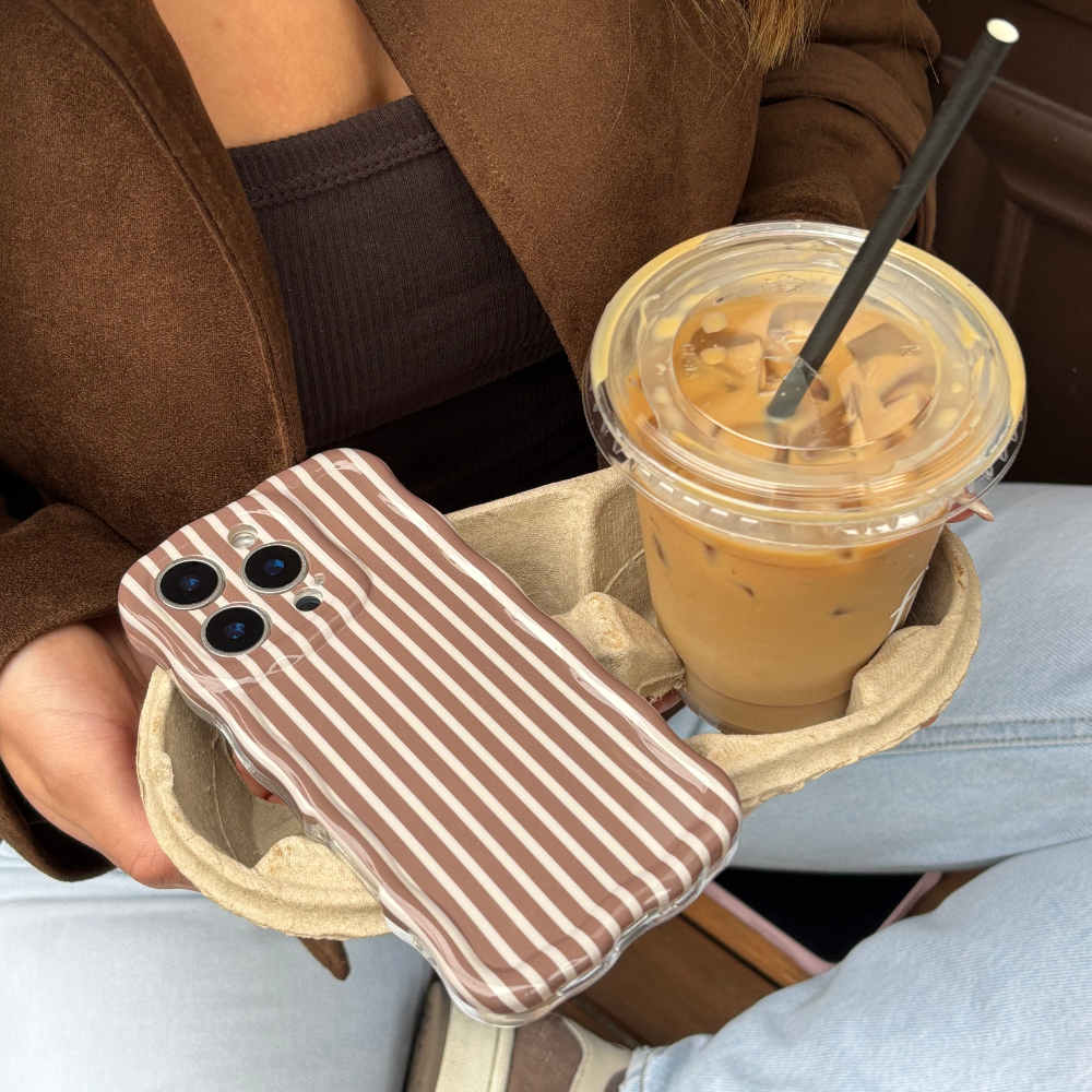 Hand holding Coconut Lane wavy phone case in chestnut stripes with iced coffee; person wears brown jacket and jeans.