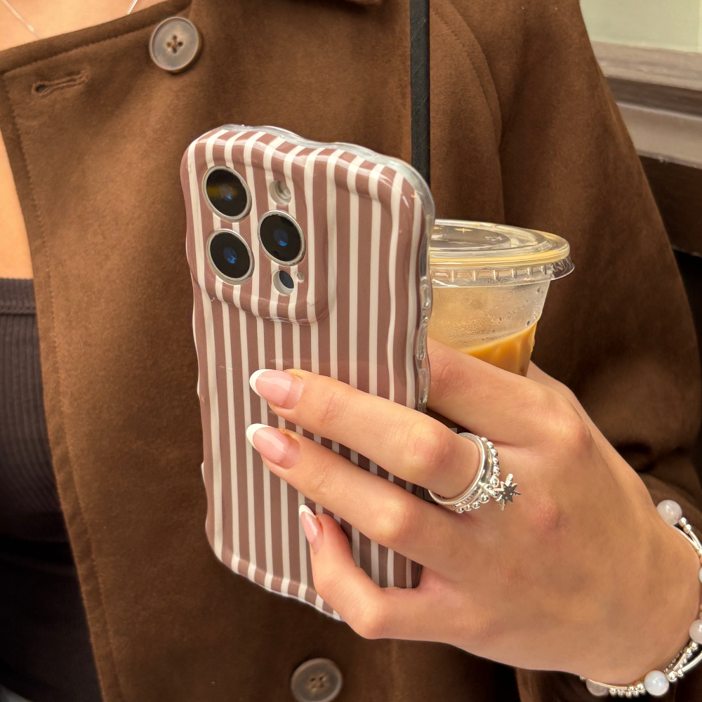 Hand with rings holds iced coffee and phone with Coconut Lane wavy chestnut striped case, brown coat in background.