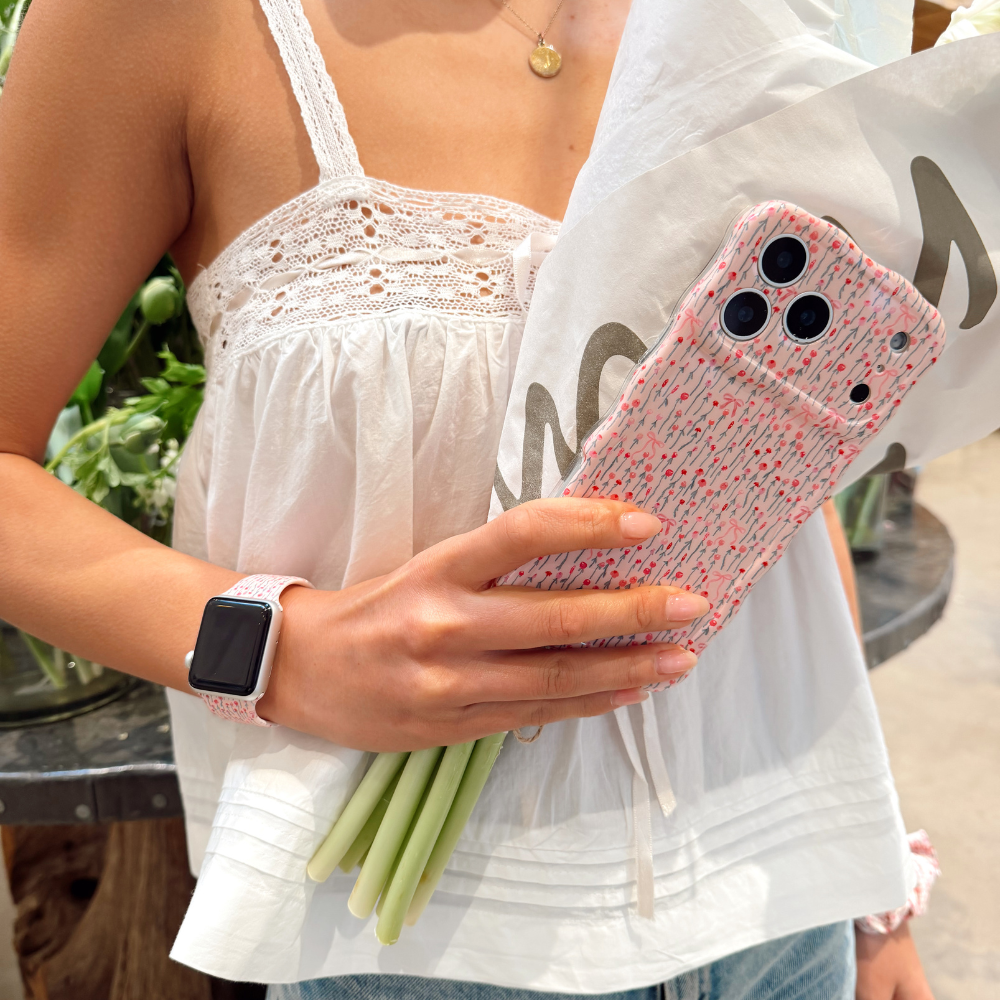 Woman in white top holds flowers and a phone with Coconut Lane pink wavy case featuring a bow design.