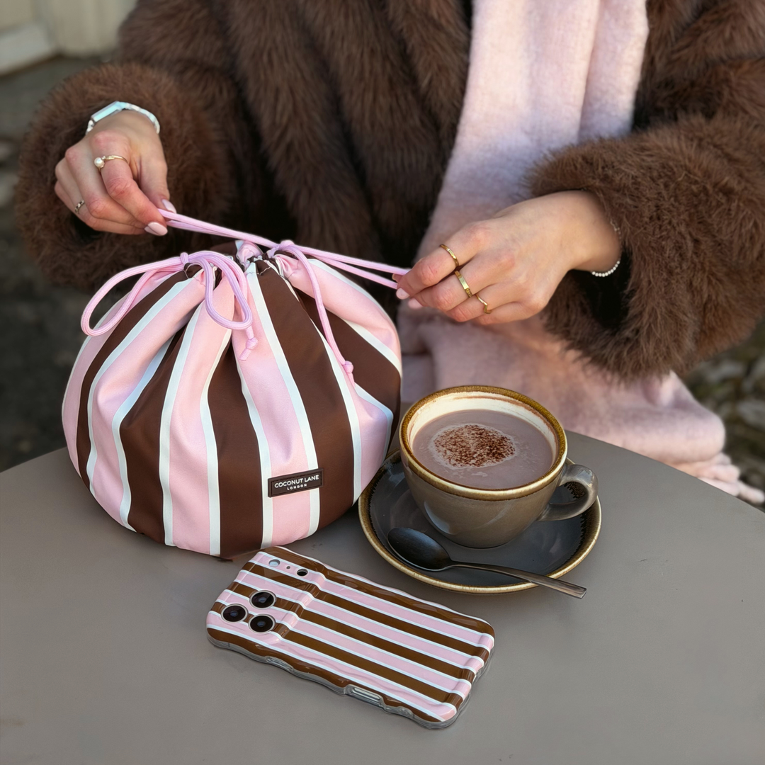 Woman wearing a baby pink scarf and brown faux fur jacket, holding a drawstring toiletry bag featuring chocolate brown, baby pink and white stripes with pink drawstring closure. Sat on the table next to this is the matching wavy phone case.