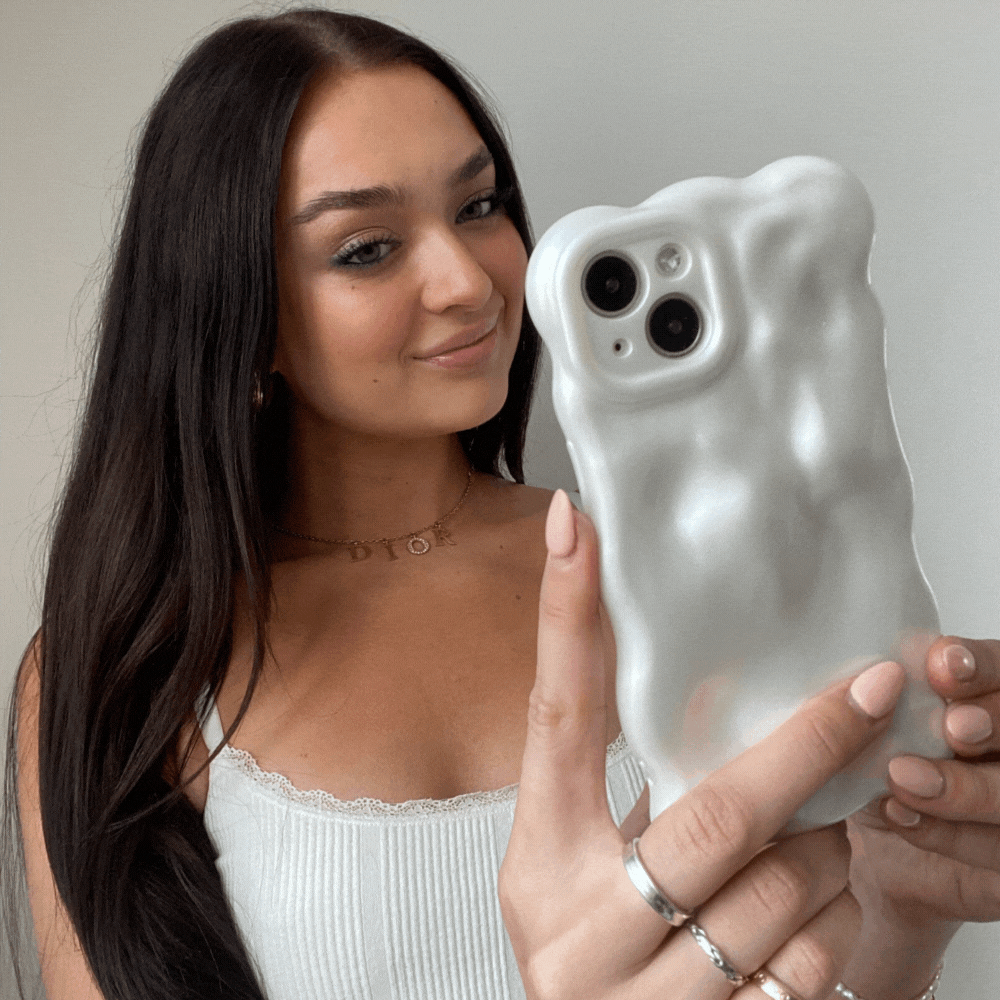 A young woman with long brown hair holds up her iPhone15 encased in Coconut Lane's 3D Pearl Phone Case - White. She is smiling and wearing a white tank top with lace trim, along with several rings and a delicate necklace. The background is plain and light-colored, emphasizing the pearl white effect of her phone case.