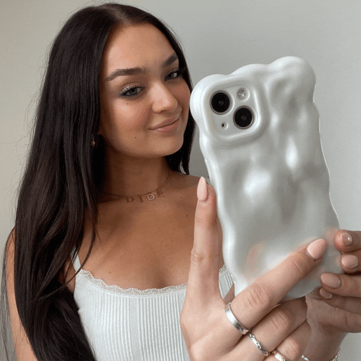 A young woman with long brown hair holds up her iPhone15 encased in Coconut Lane's 3D Pearl Phone Case - White. She is smiling and wearing a white tank top with lace trim, along with several rings and a delicate necklace. The background is plain and light-colored, emphasizing the pearl white effect of her phone case.