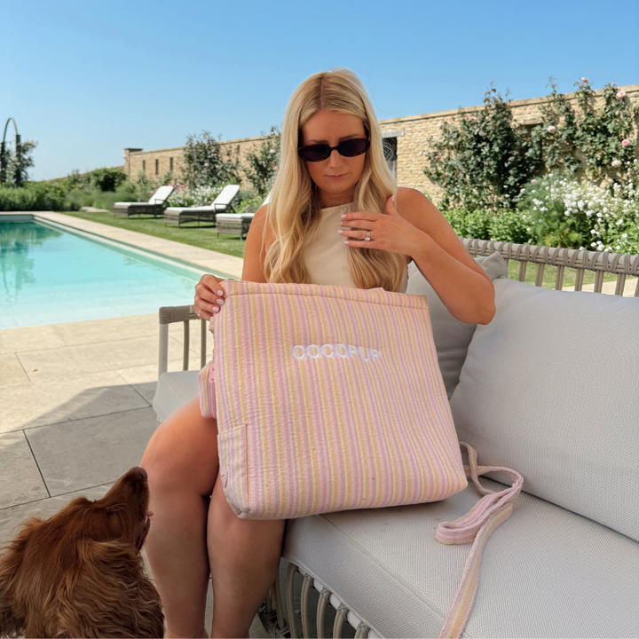 Woman by a pool with her dog holding a pastel striped tote bag with white handles.