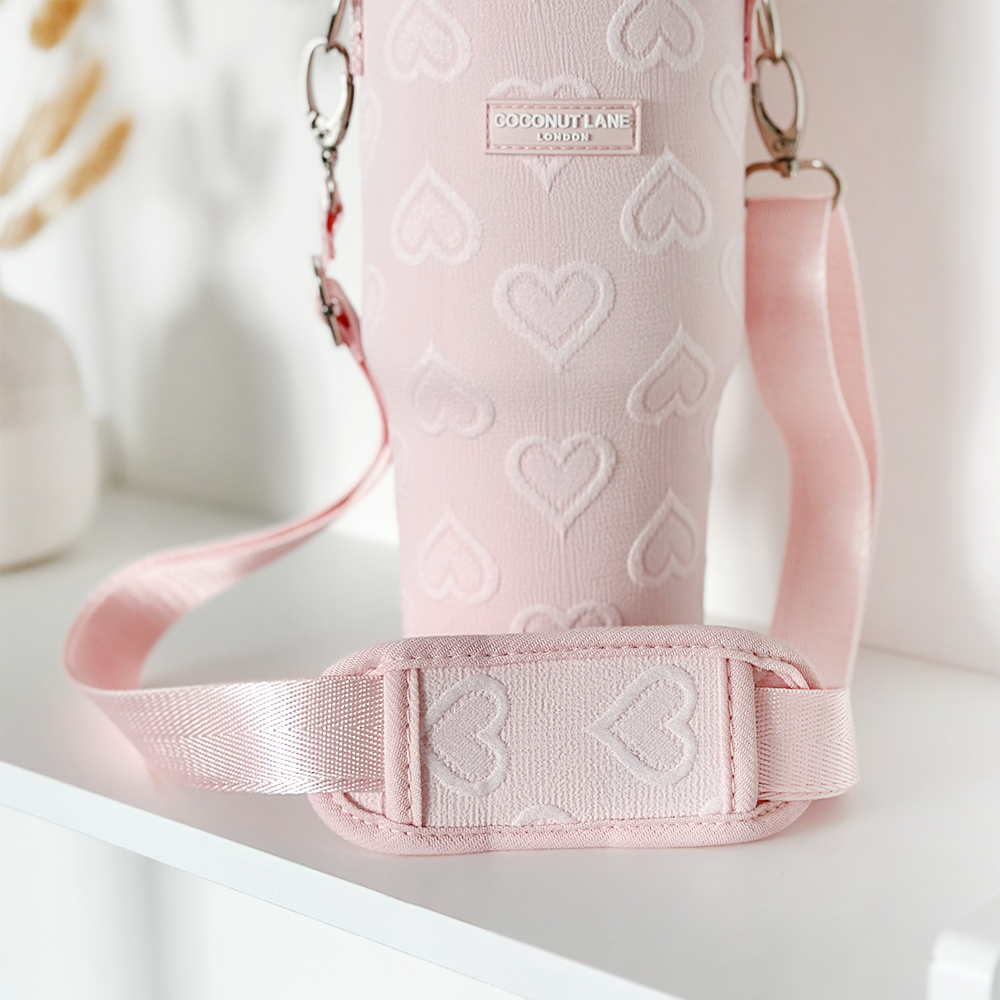 A close-up shot of the "Tumbler Carry Case - Baby Pink Heart" by Coconut Lane, featuring a charming heart pattern and an adjustable strap, placed on a white surface. The case is labeled with "Coconut Lane London." In the blurred background, minimal decor can be seen. This chic carrier provides stylish protection for your travel mug.