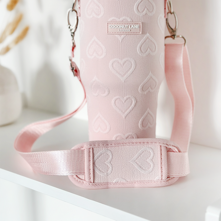 A close-up shot of the "Tumbler Carry Case - Baby Pink Heart" by Coconut Lane, featuring a charming heart pattern and an adjustable strap, placed on a white surface. The case is labeled with "Coconut Lane London." In the blurred background, minimal decor can be seen. This chic carrier provides stylish protection for your travel mug.