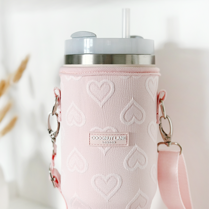 The Tumbler Carry Case - Baby Pink Heart from Coconut Lane is a pastel pink insulated cup holder decorated with heart patterns and a convenient carrying strap. It features a stitched label that reads "Coconut Lane London." The lid, made of stainless steel, has a transparent straw extending from it. The background is softly blurred.