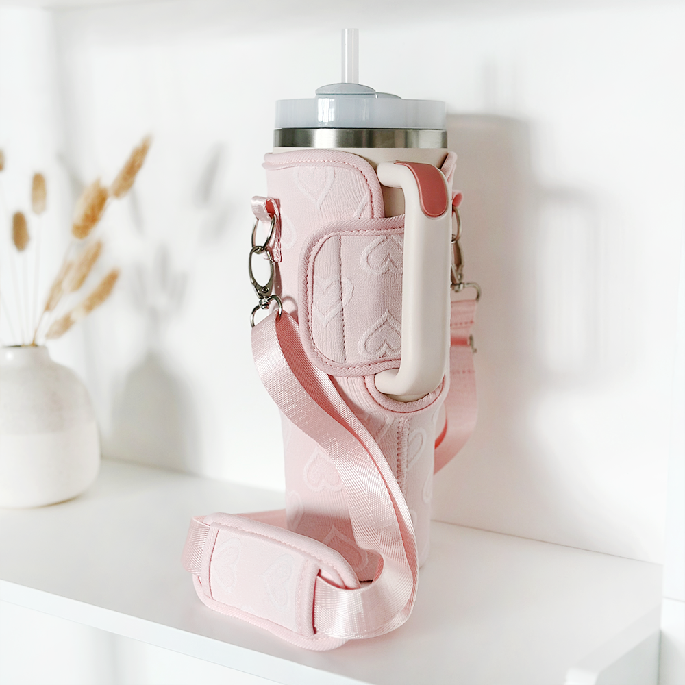 A Coconut Lane Tumbler Carry Case - Baby Pink Heart with a long strap securely holds a travel mug with a straw. The holder, adorned with handle and heart patterns, is placed on a white shelf next to a small vase containing dried wheat stalks, ensuring stylish tumbler protection.