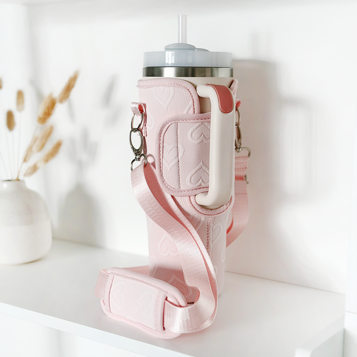 A Coconut Lane Tumbler Carry Case - Baby Pink Heart with a long strap securely holds a travel mug with a straw. The holder, adorned with handle and heart patterns, is placed on a white shelf next to a small vase containing dried wheat stalks, ensuring stylish tumbler protection.