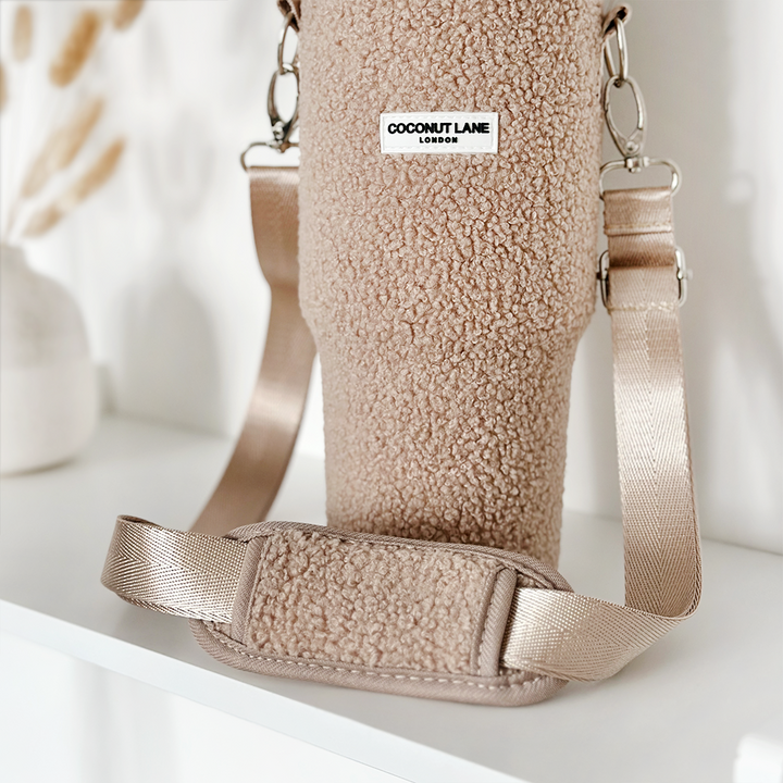 A Teddy Rupert Tumbler Carry Case from Coconut Lane, featuring a metallic beige strap, sits on a white shelf. This fuzzy beige holder is ideal for travel mug protection and has a label reading "Coconut Lane London." The background showcases a blurred white vase with dried beige plants.