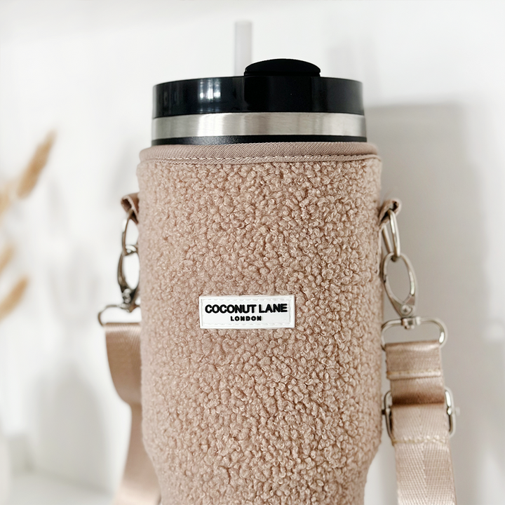 The Tumbler Carry Case - Teddy Rupert from Coconut Lane includes a reusable tumbler with a black lid and straw, encased in a beige, fuzzy holder that proudly displays the "Coconut Lane London" label. It offers added convenience with its detachable strap and metal clasps. The background is a blurred, light-colored setting.