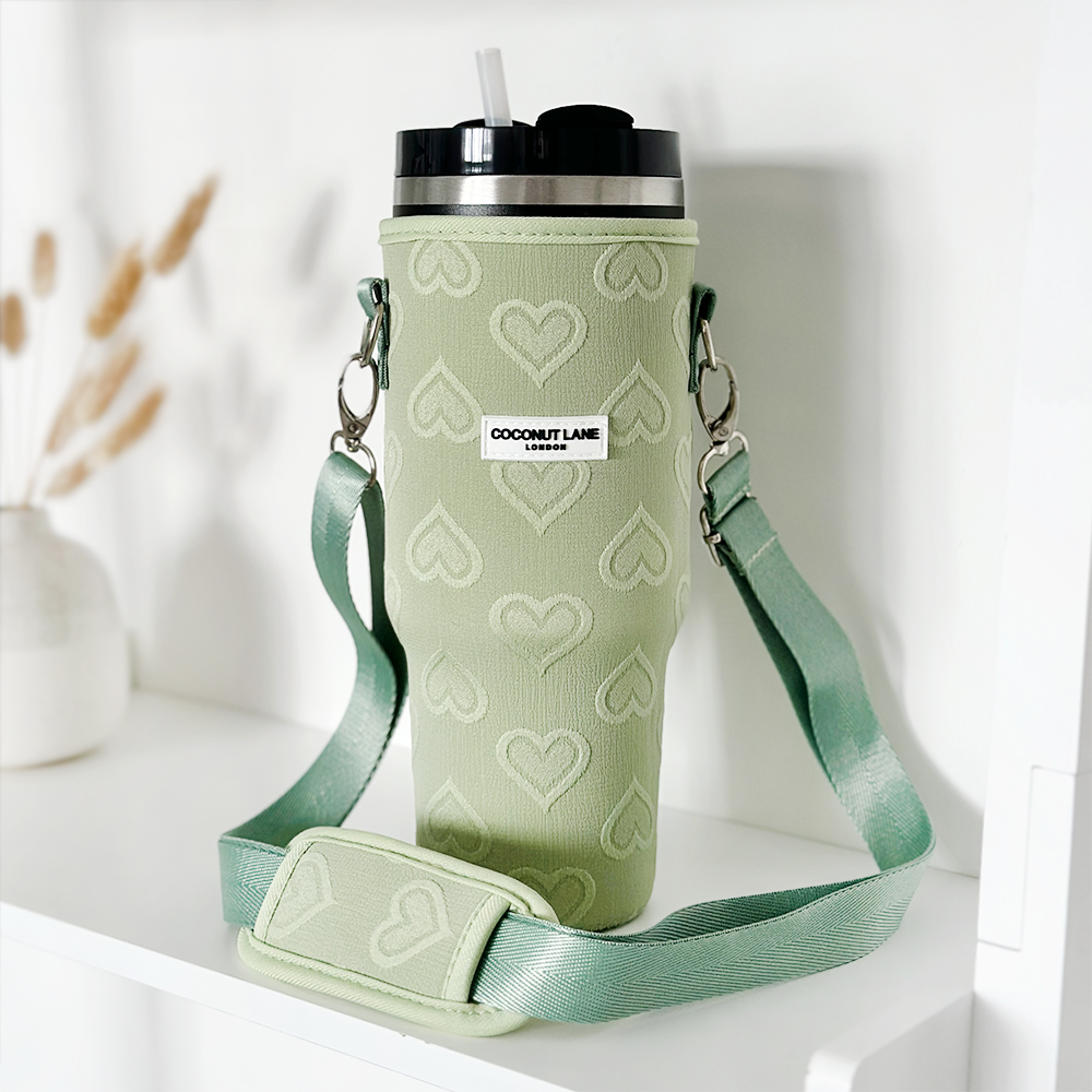 A green insulated Stanley Quencher tumbler, featuring a black lid and white straw, is showcased alongside a matching "Tumbler Carry Case - Sage Heart." The travel mug bag from Coconut Lane is decorated with heart patterns and includes a shoulder strap with the "Coconut Lane" label. In the background, dried plants and a white vase are visible.