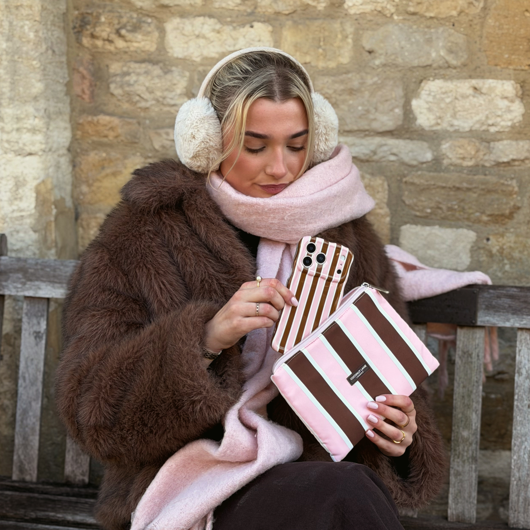 Woman wearing a baby pink scarf and faux fur brown jacket with cream fluffy ear muffs on her head. She is holding an iphone in a cocoa & Pink Stripe Wavy Phone Case featuring glossy brown, blush pink and cream vertical stripes, photographed diagonally on a pink background. In her hand is the matching Cocoa and Pink stripes everything pouch. 