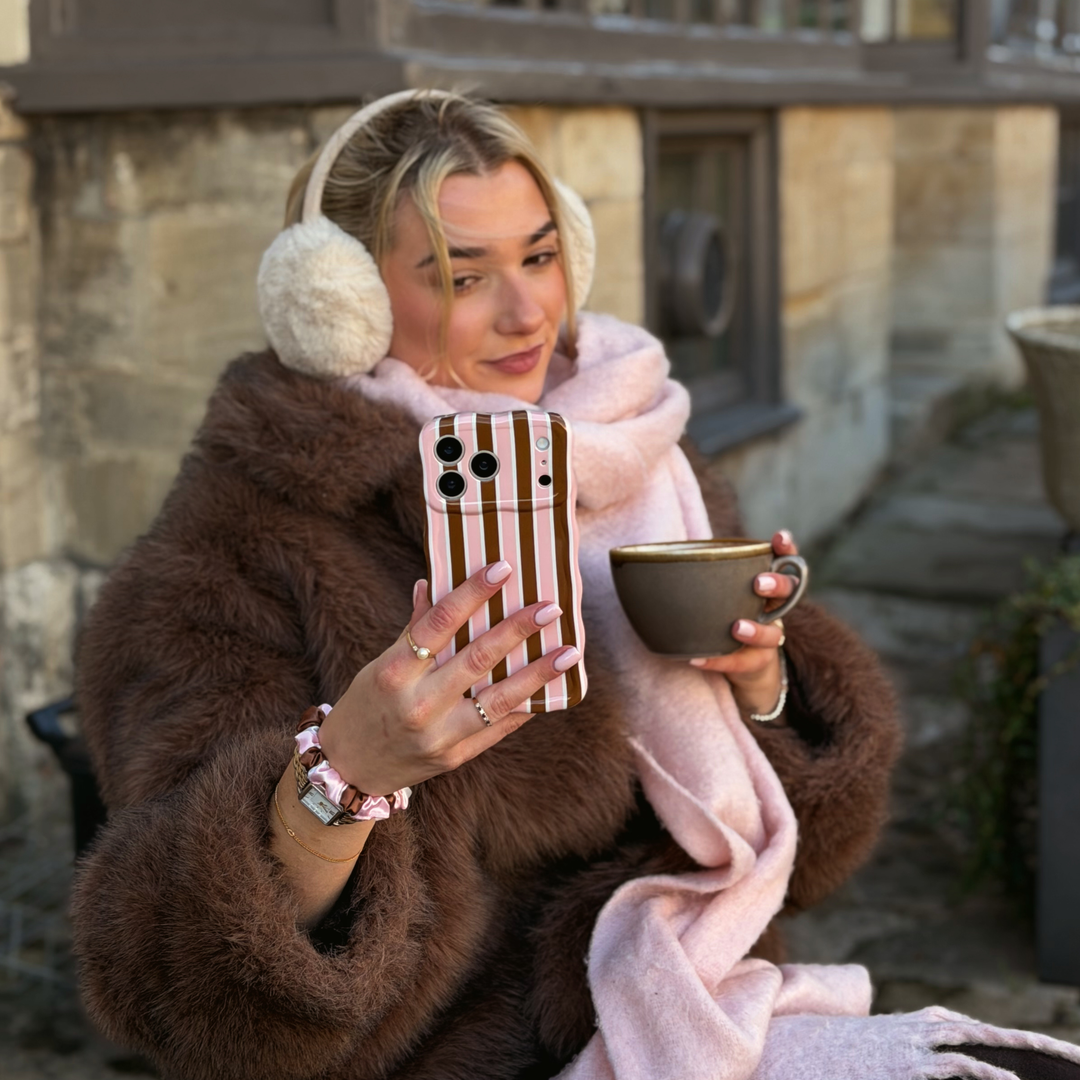 Woman taking a selfie with an iphone in a cocoa & Pink Stripe Wavy Phone Case featuring glossy brown, blush pink and cream vertical stripes, photographed diagonally on a pink background. She is wearing a baby pink scarf and faux fur brown jacket with cream fluffy ear muffs on her head. 