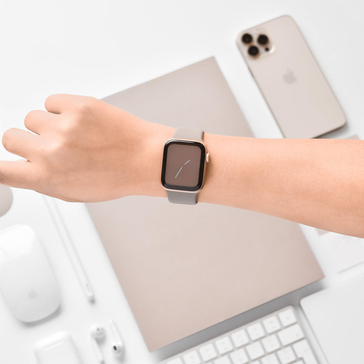 An arm wearing a NAKD Apple Watch Strap - Mocha from Coconut Lane is shown over a flat lay of Apple products, including an iPhone, a MacBook, a mouse, a keyboard, a pencil, and earphones from the NAKD Collection. All devices are arranged on a white surface, emphasizing the sleek and tech-savvy setup.