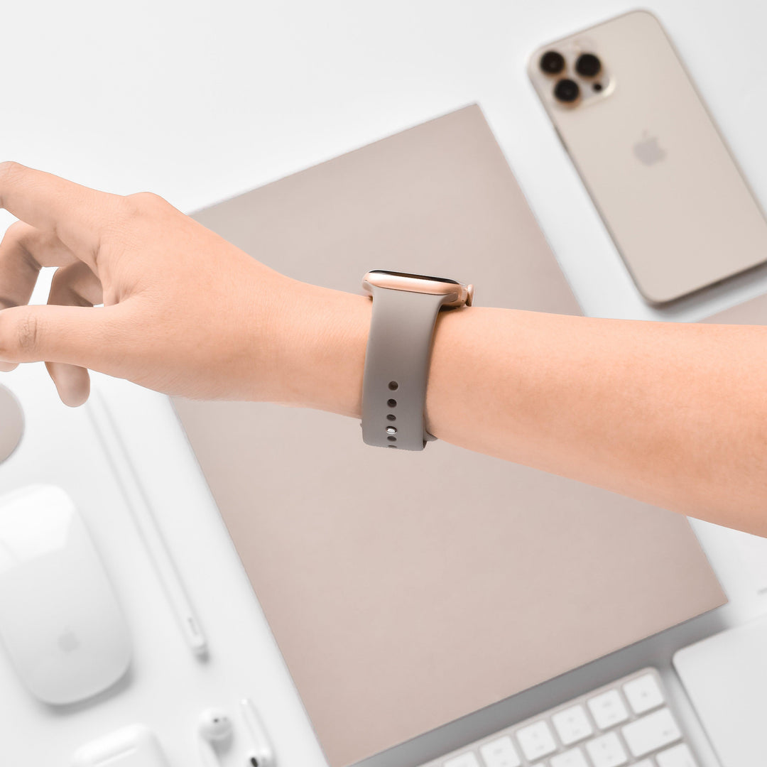 A wrist adorned with a NAKD Apple Watch Strap - Mocha by Coconut Lane is held above a workspace. On the desk are a smartphone, a closed laptop, a keyboard, a mouse, a mouse pad, and several pens, all in neutral and white tones from the NAKD Collection.