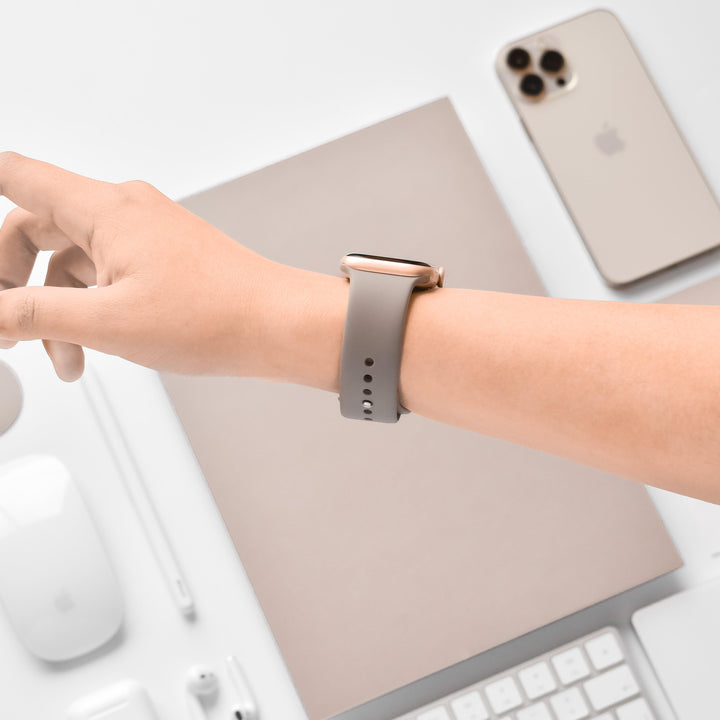 A wrist adorned with a NAKD Apple Watch Strap - Mocha by Coconut Lane is held above a workspace. On the desk are a smartphone, a closed laptop, a keyboard, a mouse, a mouse pad, and several pens, all in neutral and white tones from the NAKD Collection.