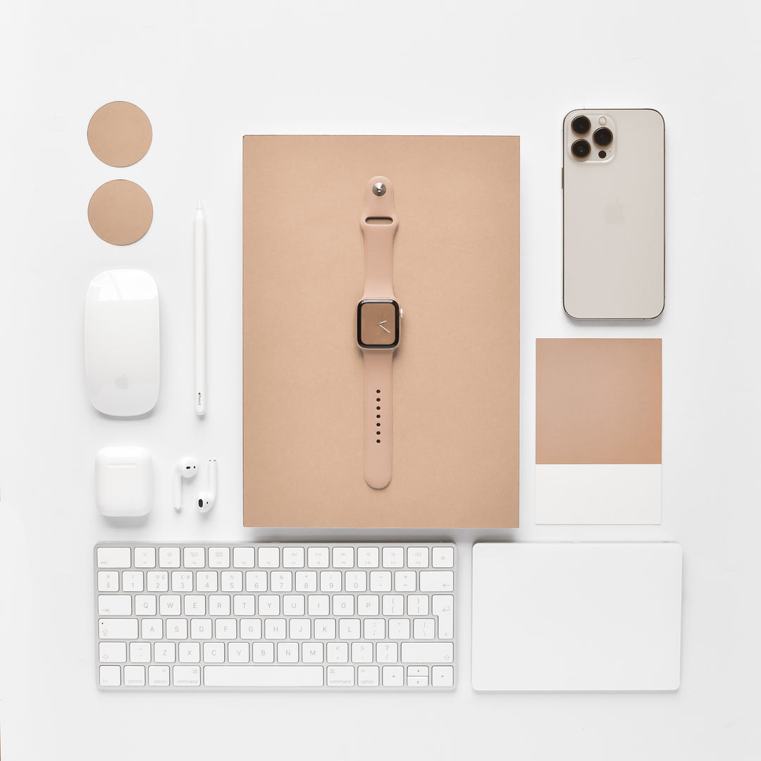 Flat lay of tech gadgets and stationery items on a white surface, featuring the NAKD Apple Watch Strap - Cappuccino by Coconut Lane centered on a beige notebook, an iPhone, a wireless mouse, a keyboard, a stylus, AirPods, and various round and square beige paper swatches from the NAKD Collection.