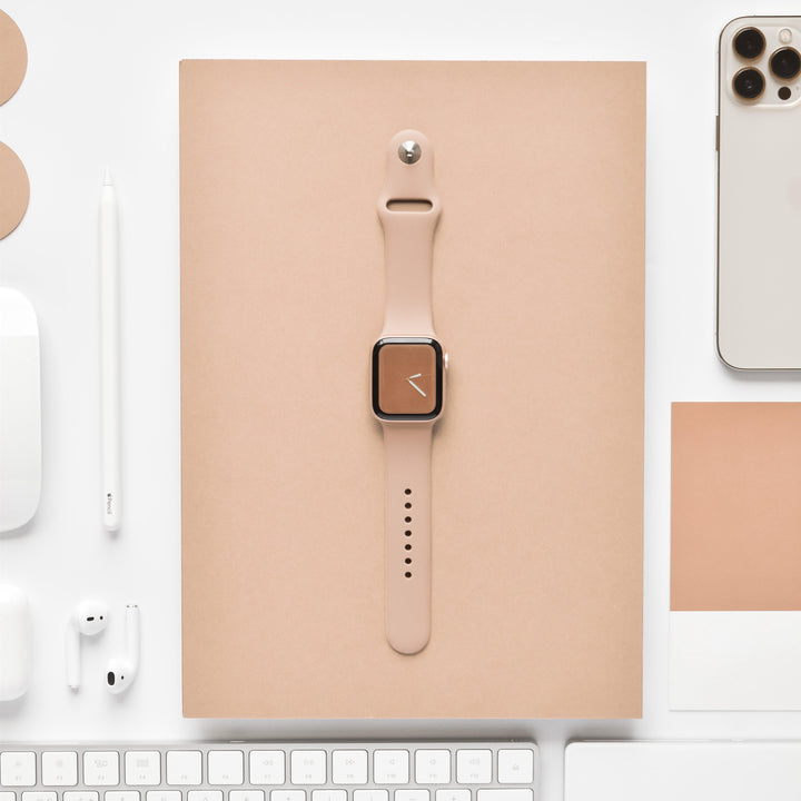 A flat lay photograph showcasing the NAKD Collection of beige and white tech and stationery items on a white background, featuring a smartwatch with the Coconut Lane NAKD Apple Watch Strap in Cappuccino, a beige notebook, an iPhone, a computer keyboard, earphones, a mouse, a pen, and color swatches.