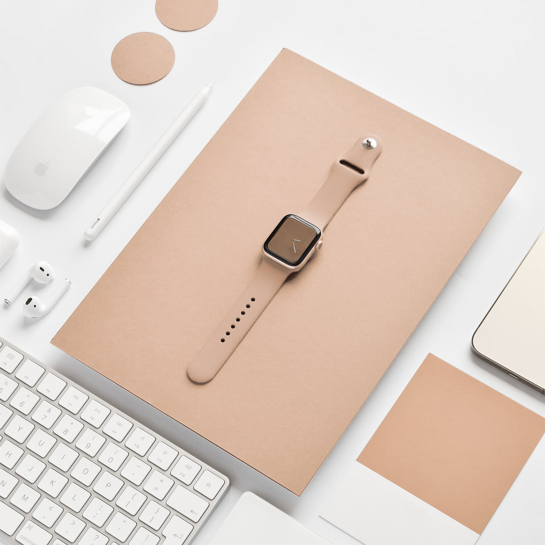 A beige smartwatch with a square face sits on a beige notebook, surrounded by an array of white office essentials such as a keyboard, mouse, pen, earphones, and laptop. Two round beige coasters are also visible. The NAKD Apple Watch Strap - Cappuccino by Coconut Lane adds an elegant touch to the setup.