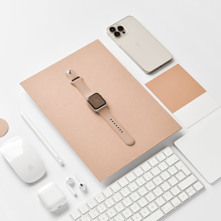 A neatly arranged workspace featuring a smartwatch with a NAKD Apple Watch Strap - Cappuccino by Coconut Lane, a beige smartphone, white wireless earbuds, a white stylus, a white computer mouse, a white keyboard, a white notebook from the NAKD Collection, and color swatches in beige tones on a beige notepad.