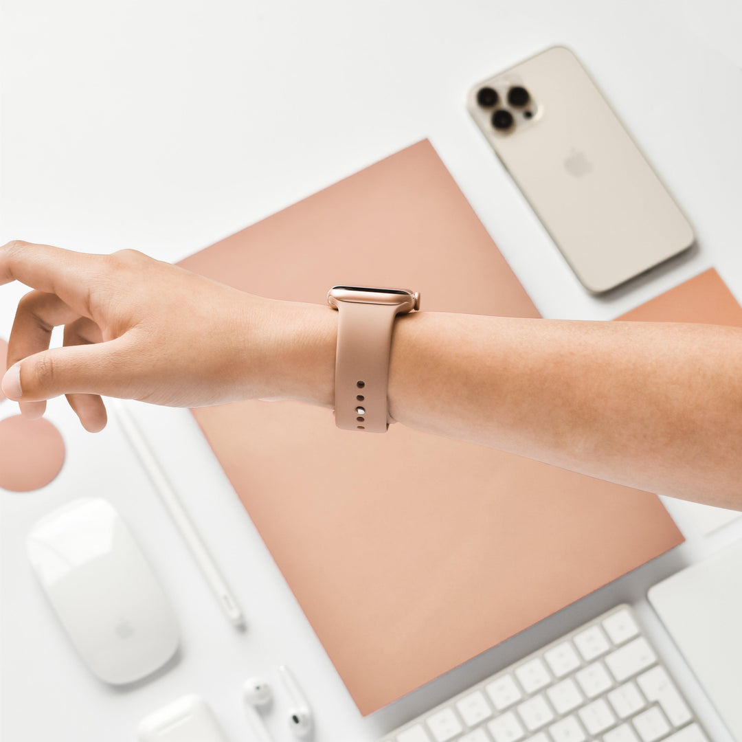A person's left arm adorned with the Coconut Lane NAKD Apple Watch Strap in Cappuccino extends over a white desk, showcasing a beige smartphone, wireless earbuds in their case, a white keyboard and mouse, a beige stylus, a circular mouse pad, and a peach-colored notebook from the NAKD Collection.