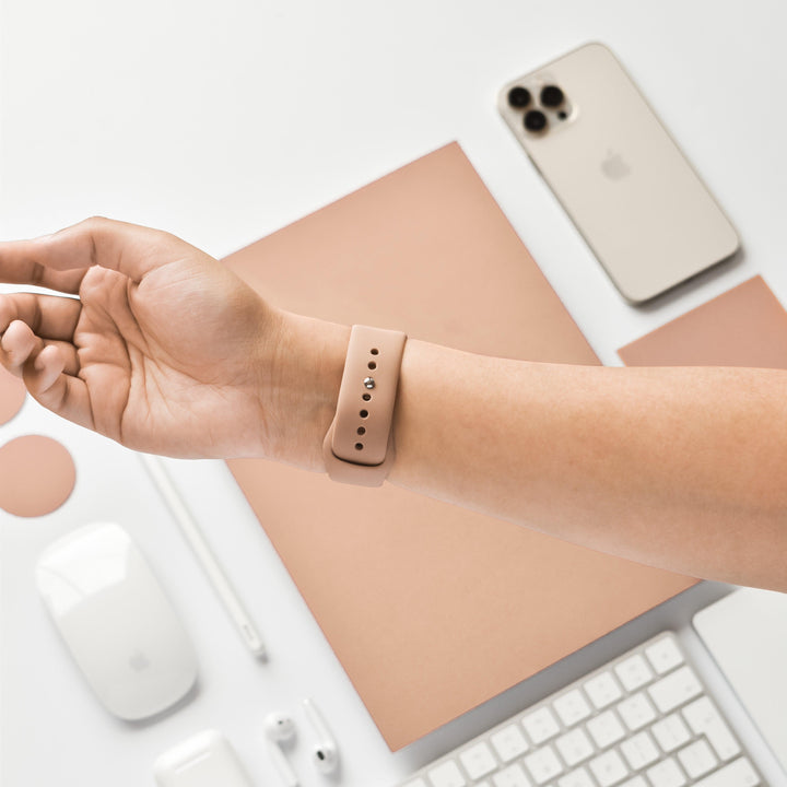 A person's wrist with a Coconut Lane NAKD Apple Watch Strap in Cappuccino is in the foreground. In the background, a flat lay displays an Apple iPhone, wireless earbuds, a mouse, a keyboard, a stylus, and a mouse pad— all in white or beige on a clean, light surface from the NAKD Collection.