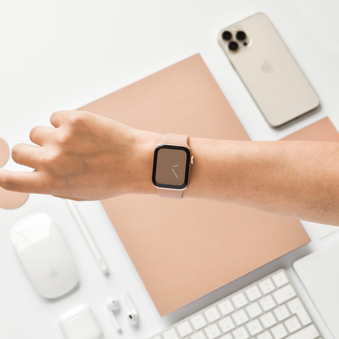 A person is wearing the Coconut Lane NAKD Apple Watch Strap in Cappuccino on their wrist. The background features an Apple iPhone, a wireless mouse, a stylus, earphones, a keyboard, and a notebook, all in beige and white tones. This minimalist tech setup is enhanced by the stylish NAKD Collection silicone Apple Watch Bands from Coconut Lane.