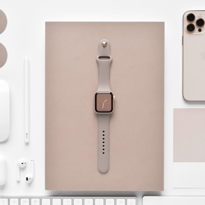 A beige smart watch adorned with the Coconut Lane NAKD Apple Watch Strap in Mocha is showcased on top of a beige notebook. Encircling the notebook are a white keyboard, a white mouse, white earphones, a white stylus, and a gold smartphone from the NAKD Collection, all meticulously arranged on an immaculate white surface.