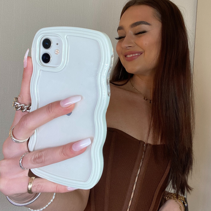A young woman with long brown hair smiles while taking a selfie. She wears a brown, strapless top and has multiple rings on her fingers. Her iPhone case by Coconut Lane, the Curvy Phone Case in Cream, notably a highly protective design with sleek curves, stands out against the plain background.