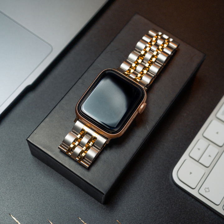 A smartwatch featuring the Stainless Steel Apple Watch Strap in Silver & Gold from Coconut Lane is placed on a black box. The watch screen appears blank and reflective. The box rests on a dark surface, surrounded by parts of a laptop and a white keyboard.
