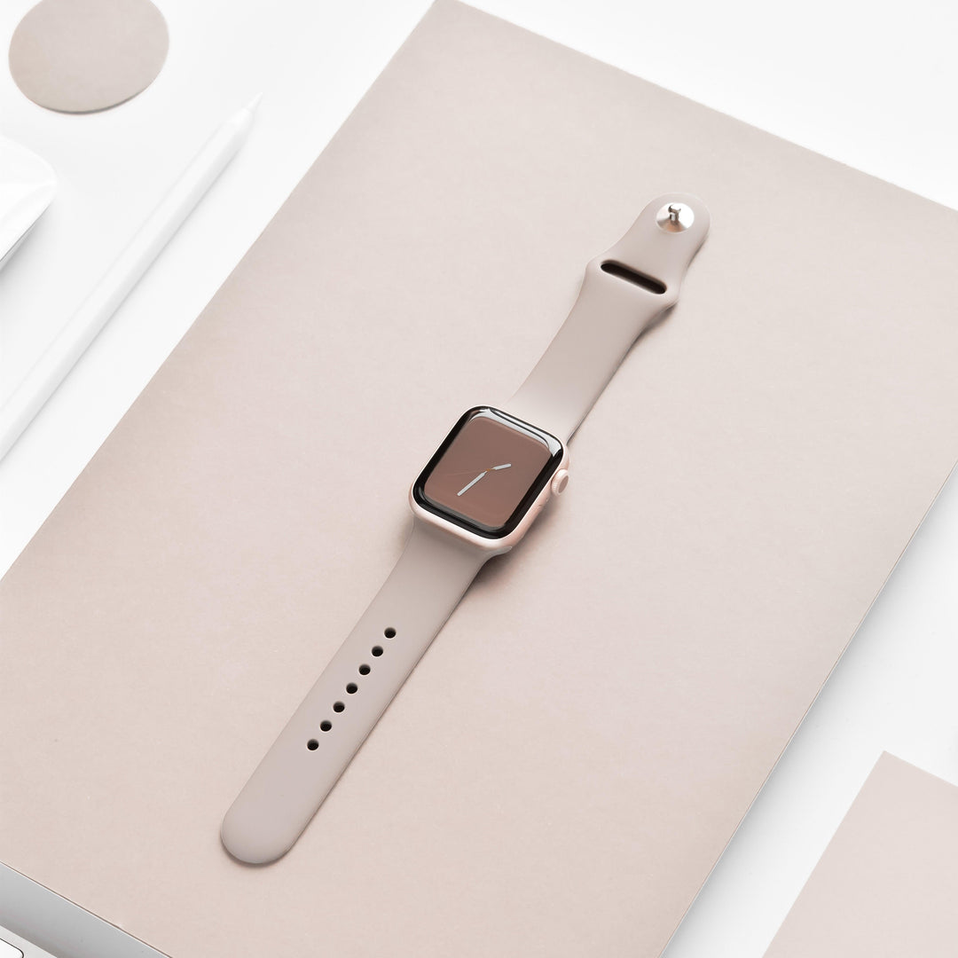A NAKD Apple Watch Strap - Mocha from Coconut Lane, featuring a beige silicone band and a rectangular face displaying a minimalist clock, is placed on a beige surface. Surrounding the watch are various minimalistic items, including a white pen, a small white object, and the corner of a white screen.