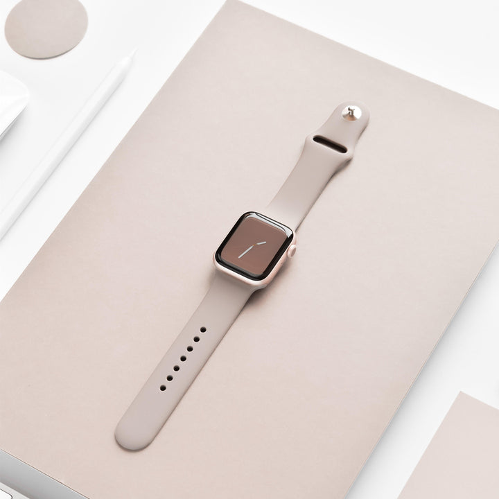 A NAKD Apple Watch Strap - Mocha from Coconut Lane, featuring a beige silicone band and a rectangular face displaying a minimalist clock, is placed on a beige surface. Surrounding the watch are various minimalistic items, including a white pen, a small white object, and the corner of a white screen.