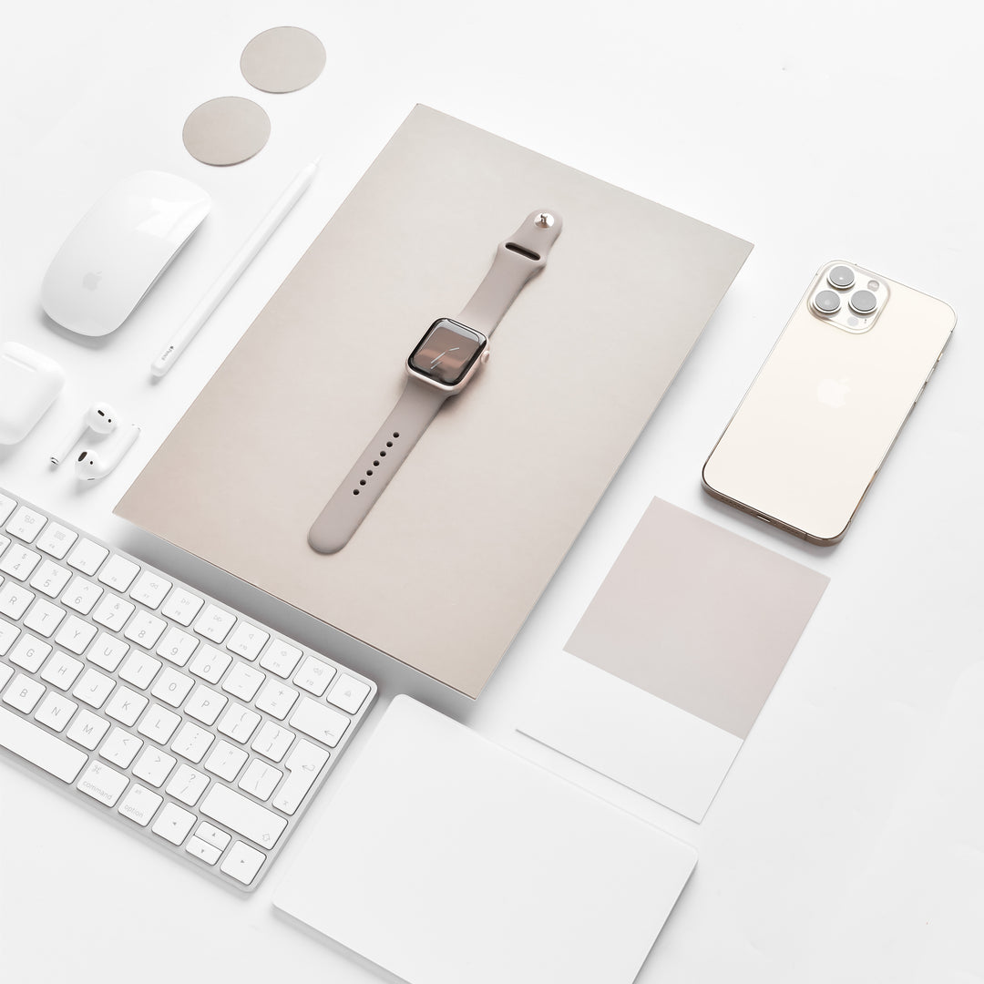 A neatly arranged flat lay features a tablet with a NAKD Apple Watch Strap - Mocha by Coconut Lane, a smartphone, a keyboard, a wireless mouse, earbuds, a stylus, circular and square swatches from the NAKD Collection, and note cards. All items are primarily white or beige, creating a minimalist aesthetic.