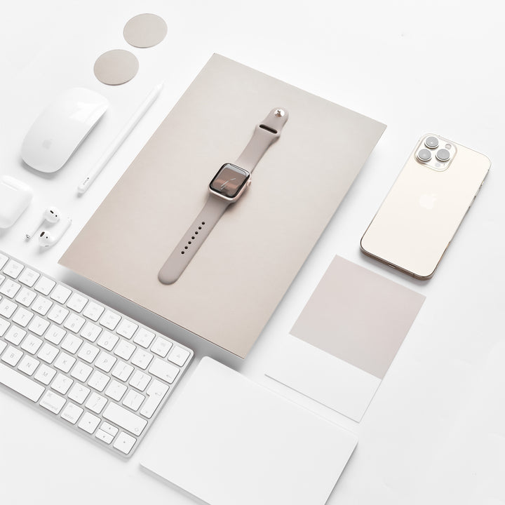 A neatly arranged flat lay features a tablet with a NAKD Apple Watch Strap - Mocha by Coconut Lane, a smartphone, a keyboard, a wireless mouse, earbuds, a stylus, circular and square swatches from the NAKD Collection, and note cards. All items are primarily white or beige, creating a minimalist aesthetic.