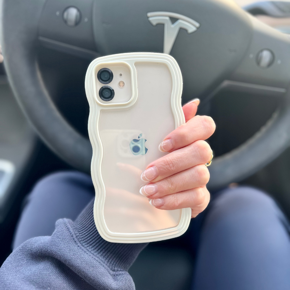 A person with manicured nails holds an iPhone encased in the highly protective Curvy Phone Case - Cream by Coconut Lane, featuring a wavy design. The steering wheel adorned with the Tesla logo is visible in the background, indicating they are seated in the driver's seat of a Tesla vehicle.
