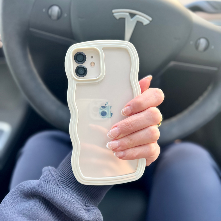 A person with manicured nails holds an iPhone encased in the highly protective Curvy Phone Case - Cream by Coconut Lane, featuring a wavy design. The steering wheel adorned with the Tesla logo is visible in the background, indicating they are seated in the driver's seat of a Tesla vehicle.