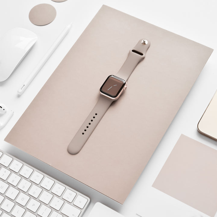 A rose gold smartwatch with a NAKD Apple Watch Strap - Mocha from Coconut Lane sits on top of a beige notebook. Surrounding it are white tech accessories like a keyboard, stylus, computer mouse, and earphones, all from the NAKD Collection, neatly arranged on a white surface.