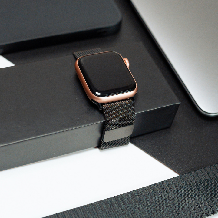 A Coconut Lane smartwatch featuring a Magnetic Chain Apple Watch Strap in black and a casing in rose gold is displayed atop a black rectangular box. The background showcases parts of a laptop and other electronic devices, all set in an elegant, modern environment.