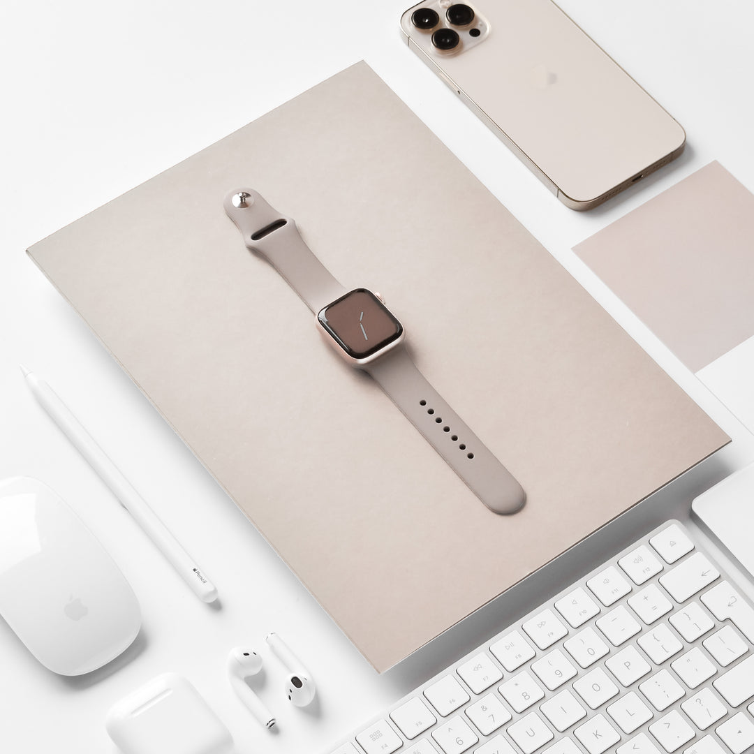A beige Apple Watch with the Coconut Lane NAKD Apple Watch Strap - Mocha is displayed on top of a beige document or booklet. Surrounding it are a gold iPhone, a white Apple Magic Mouse, AirPods, an Apple Pencil, and a white keyboard, all arranged on a pristine white desk.