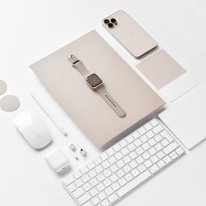 A neatly arranged display of Apple products including an iPhone, an Apple Watch with the NAKD Mocha strap by Coconut Lane, AirPods, a wireless keyboard, a Magic Mouse, and an Apple Pencil on a white desk. The items are organized in a visually pleasing layout with the Apple Watch displayed centrally on a beige booklet.