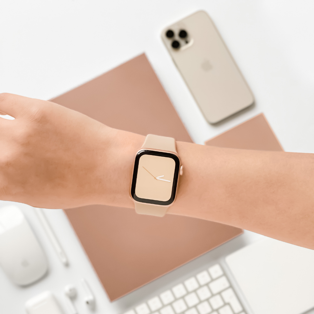 A person's wrist is shown wearing a beige smartwatch with the NAKD Apple Watch Strap in Caramel Latte from Coconut Lane, which complements its minimalist design. In the background, there is a beige smartphone, a beige laptop, and various accessories including a mouse, keyboard, and cables, all neatly arranged on a light surface.