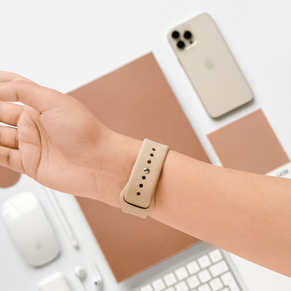 A person's wrist is shown wearing a beige smartwatch with the Coconut Lane NAKD Apple Watch Strap in Caramel Latte. The wrist is positioned above a minimalistic workspace that includes a beige smartphone, a mouse, a keyboard, a pen, and a brown notebook all laid out on a white surface.