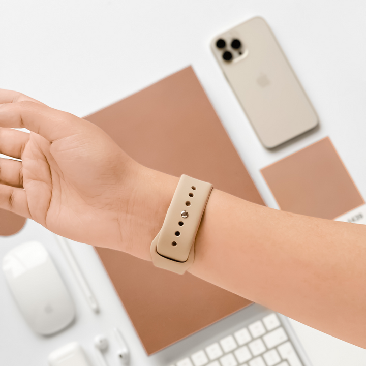 A person's wrist is shown wearing a beige smartwatch with the Coconut Lane NAKD Apple Watch Strap in Caramel Latte. The wrist is positioned above a minimalistic workspace that includes a beige smartphone, a mouse, a keyboard, a pen, and a brown notebook all laid out on a white surface.