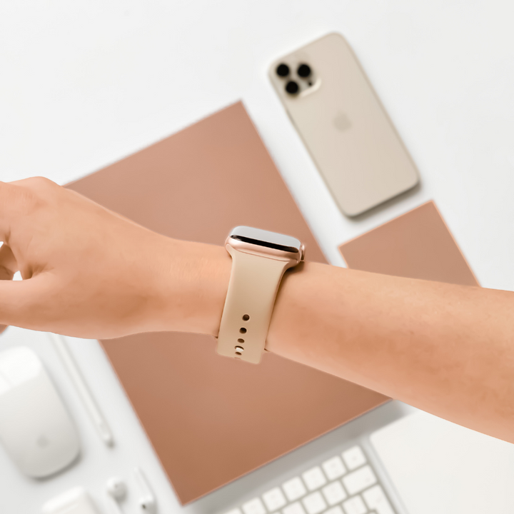 Close-up of an outstretched arm wearing a beige-colored smartwatch with the stylish NAKD Apple Watch Strap - Caramel Latte by Coconut Lane. In the background, there are a smartphone, a laptop, a tablet, and other office accessories in matching beige and white tones on a pristine white surface.