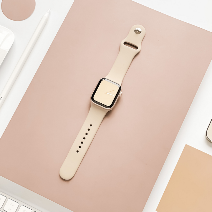 A beige smartwatch with a matching Coconut Lane NAKD Apple Watch Strap - Caramel Latte is placed on a pinkish-beige surface, surrounded by a white pencil, part of a keyboard, and a square sample in a similar hue. The display shows a minimalist clock face design from the NAKD Collection.