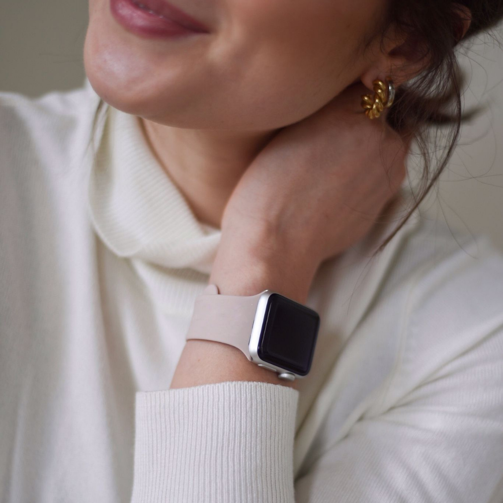 Close-up of a person wearing a dusty pink, durable and chic NAKD Apple Watch Strap by Coconut Lane on their wrist, partially concealing their face. They are smiling softly, wearing a cream-colored sweater and gold floral earrings. The background is blurred and neutral-toned.