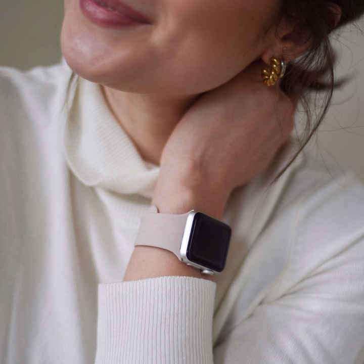 Close-up of a person wearing a dusty pink, durable and chic NAKD Apple Watch Strap by Coconut Lane on their wrist, partially concealing their face. They are smiling softly, wearing a cream-colored sweater and gold floral earrings. The background is blurred and neutral-toned.