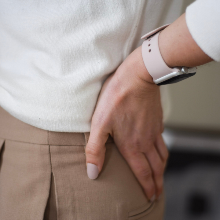 A person wearing a white shirt and beige pants is holding their lower back with their left hand, indicating back pain. They are wearing a Coconut Lane NAKD Apple Watch Strap in Dusty Pink on their wrist, and their neatly groomed nails match the color of the durable and chic silicone strap.